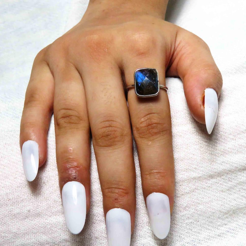 May include: A silver ring with a square, iridescent blue gemstone. The ring is on a hand with long, white fingernails. The gemstone has flashes of blue and green.