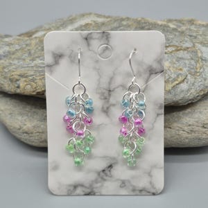May include: Dangle earrings with silver-tone metal chains and hooks. Each earring has clusters of blue, pink, and green beads. The earrings are displayed on a white marble-patterned card. The earrings are a stylish accessory.