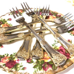 May include: Set of six gold-plated forks with rose design on the handles. The forks are arranged on a white and gold floral patterned plate.