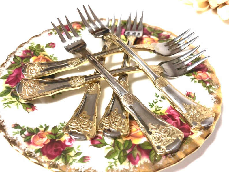 May include: Set of six gold-plated forks with rose design on the handles. The forks are arranged on a white and gold floral patterned plate.