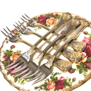 May include: Set of six silver forks with gold accents and floral designs on the handles. The forks are arranged on a white plate with a floral pattern.
