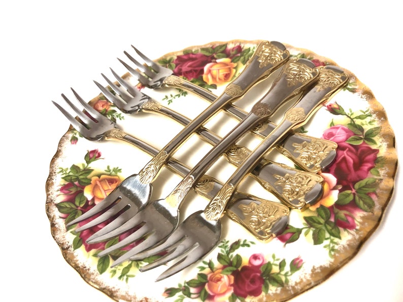 May include: Set of six silver forks with gold accents and floral designs on the handles. The forks are arranged on a white plate with a floral pattern.