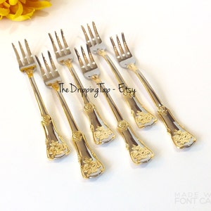May include: Set of six gold and silver dessert forks with a rose design on the handle. The forks are arranged in a row on a white surface. The text "The Dripping Tap - Etsy" is visible in the image.