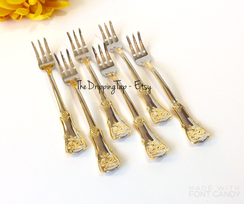 May include: Set of six gold and silver dessert forks with a rose design on the handle. The forks are arranged in a row on a white surface. The text "The Dripping Tap - Etsy" is visible in the image.