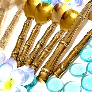 Puede incluir: Conjunto de cinco cucharas doradas de estilo bamb&uacute; con cuencos redondeados. Las cucharas est&aacute;n dispuestas sobre una superficie blanca con piedras de vidrio azules.