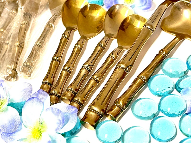 Puede incluir: Conjunto de cinco cucharas doradas de estilo bamb&uacute; con cuencos redondeados. Las cucharas est&aacute;n dispuestas sobre una superficie blanca con piedras de vidrio azules.