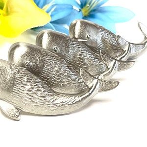 May include: A set of five silver whale figurines, arranged in a line, with the largest whale at the front and the smallest whale at the back. The whales are all facing the same direction, with their tails pointing towards the back of the image.