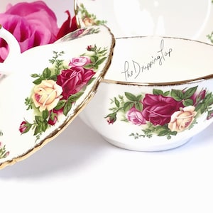 May include: A white porcelain bowl with a lid, both decorated with a floral pattern of red, pink, and yellow roses. The bowl has a gold rim and the lid has a handle in the shape of a swan. The text "The Dripping Tap" is written on the bowl.