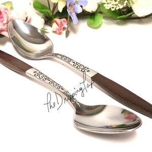 May include: Two silver spoons with ornate designs on the handles and brown wooden handles. The spoons are lying on a white surface.