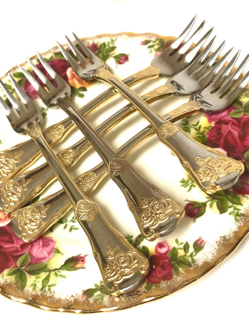 May include: Set of six gold-plated forks with rose design on a white plate with red roses.