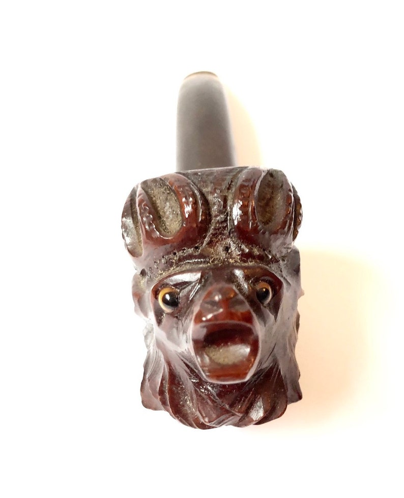 Puede incluir: Una pipa de madera marr&oacute;n oscuro tallada en forma de cabeza de animal con ojos grandes y boca abierta. La pipa es una pipa de fumar tradicional.