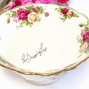 May include: A white ceramic bowl with a gold rim and a floral pattern featuring red and yellow roses. The bowl has a scalloped edge and a pedestal base.