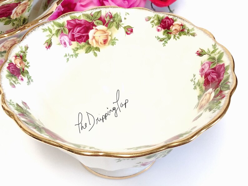 May include: A white ceramic bowl with a gold rim and a floral pattern featuring red and yellow roses. The bowl has a scalloped edge and a pedestal base.