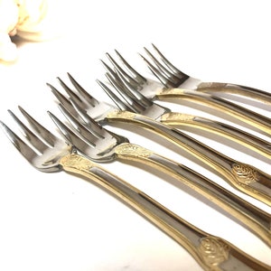 May include: Set of six gold-plated forks with ornate rose designs on the handles. The forks are made of stainless steel and have a polished finish.