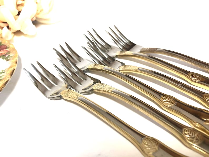 May include: Set of six gold-plated forks with ornate rose designs on the handles. The forks are made of stainless steel and have a polished finish.