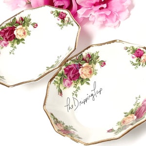 May include: Two white, octagonal serving dishes with a floral rose pattern in shades of red, pink, and yellow, accented with green leaves. The rims are trimmed with gold. The text "the Dripping Tap" is written on one dish.