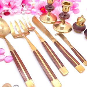 May include: A set of gold-toned serving utensils with wooden handles. The set includes a serving spoon, a serving fork, a butter knife, a cheese knife, and a small fork. The utensils are arranged on a white surface with pink flowers and decorative stones.