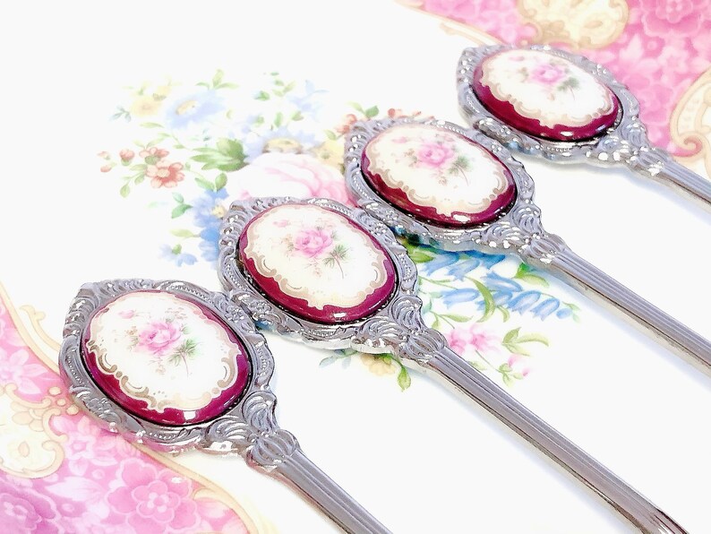 Pu&ograve; includere: Quattro cucchiaini d'argento con manici decorati e disegni floreali sulla parte superiore. I disegni floreali sono in tonalit&agrave; di rosa, bianco e oro.