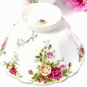 May include: A white ceramic bowl with a scalloped edge and a floral design featuring pink, yellow, and red roses. The bowl has a gold trim around the edge and a small inscription on the bottom that reads "The Romance of a Rose".