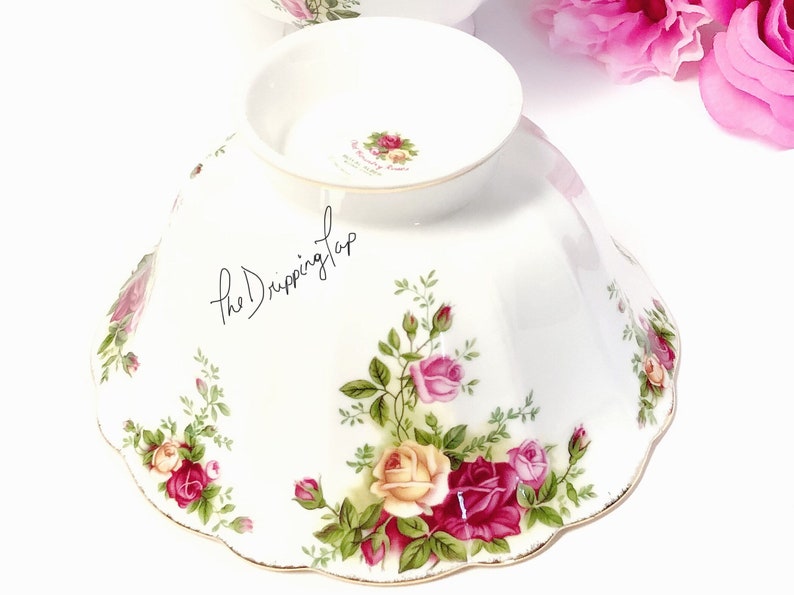 May include: A white ceramic bowl with a scalloped edge and a floral design featuring pink, yellow, and red roses. The bowl has a gold trim around the edge and a small inscription on the bottom that reads "The Romance of a Rose".