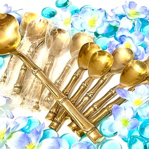 Puede incluir: Un juego de cucharas doradas con mangos de estilo bamb&uacute;. Las cucharas est&aacute;n dispuestas en forma de abanico sobre una superficie blanca con flores azules y blancas y cuentas de vidrio azules.