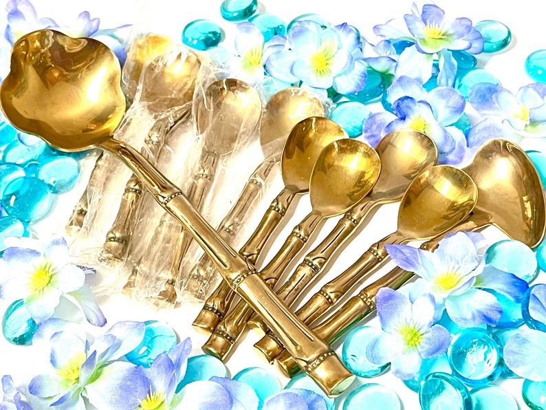 Puede incluir: Un juego de cucharas doradas con mangos de estilo bamb&uacute;. Las cucharas est&aacute;n dispuestas en forma de abanico sobre una superficie blanca con flores azules y blancas y cuentas de vidrio azules.