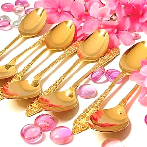 May include: Six gold-colored spoons with ornate, textured handles and polished bowls. The spoons are arranged on a white surface, surrounded by pink flowers and decorative glass beads. The spoons are designed for serving.