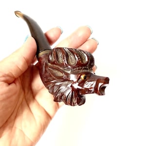 Puede incluir: Una pipa de madera tallada en forma de cabeza de lobo. La pipa es marr&oacute;n con una boquilla negra y tiene un ojo amarillo.