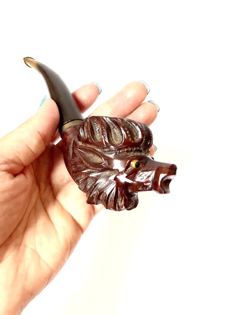 Puede incluir: Una pipa de madera tallada en forma de cabeza de lobo. La pipa es marr&oacute;n con una boquilla negra y tiene un ojo amarillo.