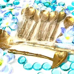 Puede incluir: Un juego de cucharas doradas con mangos de bamb&uacute;. Las cucharas est&aacute;n empaquetadas en pl&aacute;stico transparente y rodeadas de flores azules y blancas.