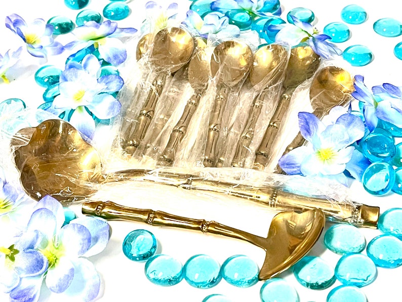 Puede incluir: Un juego de cucharas doradas con mangos de bamb&uacute;. Las cucharas est&aacute;n empaquetadas en pl&aacute;stico transparente y rodeadas de flores azules y blancas.