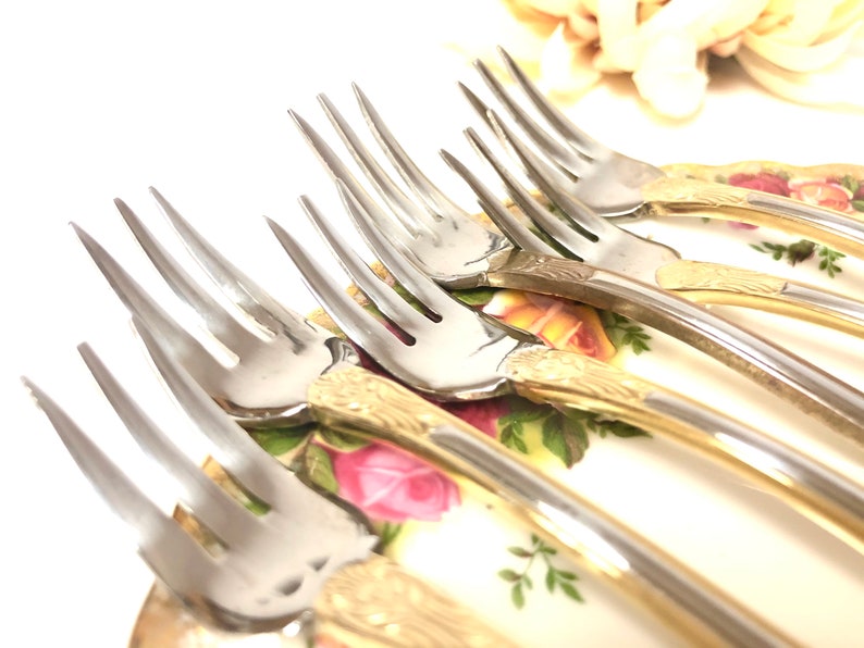 May include: A set of six silver forks with gold-plated handles. The forks are arranged in a fan shape on a floral patterned surface.