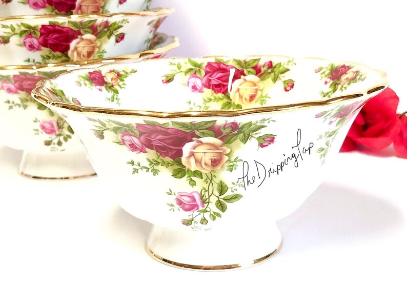 May include: A set of four white porcelain bowls with a floral pattern featuring red, pink, and yellow roses. The bowls have a gold rim and a gold trim around the base. The bowls are on a white surface. The text "the Dripping Tap" is written on the side of one of the bowls.