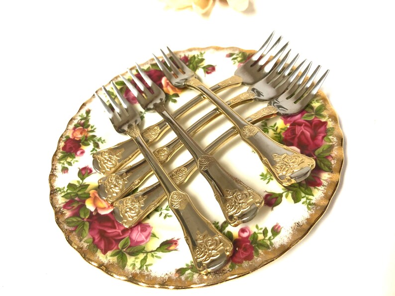 May include: Set of six gold-plated forks with ornate floral designs on a white plate with a floral pattern.