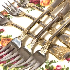 May include: Set of six silver forks with gold accents and ornate floral designs on the handles. The forks are arranged on a white plate with a floral pattern.