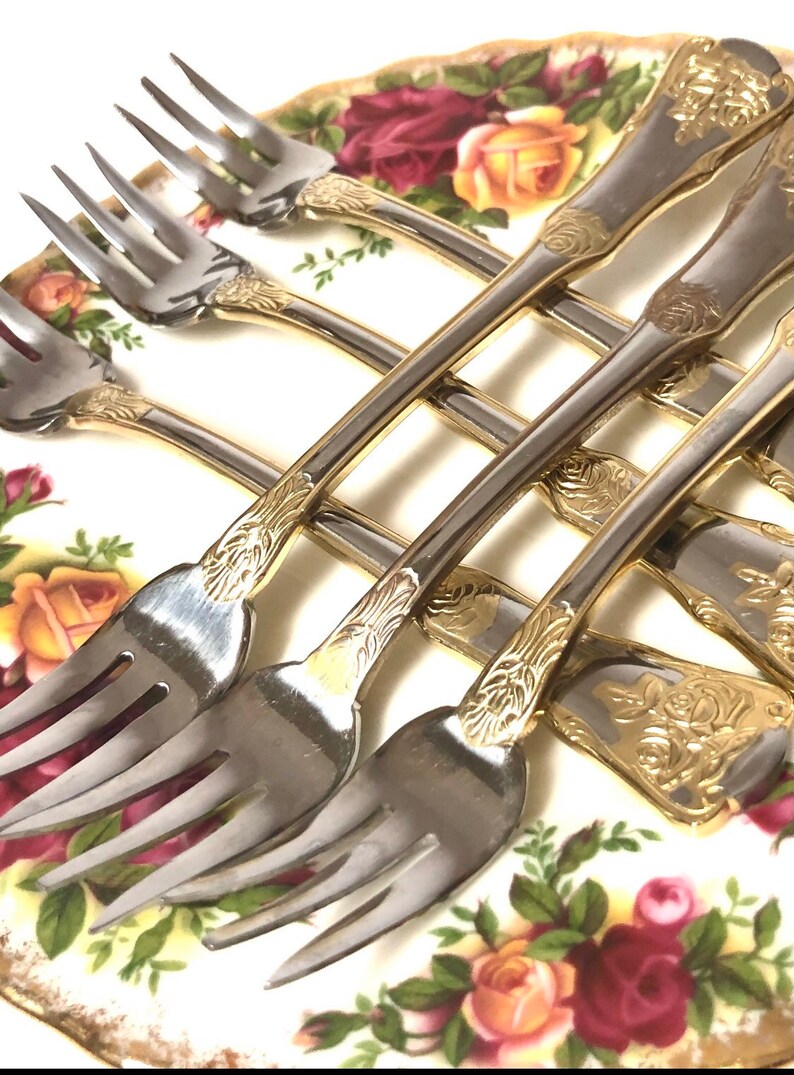 May include: Set of six silver forks with gold accents and ornate floral designs on the handles. The forks are arranged on a white plate with a floral pattern.