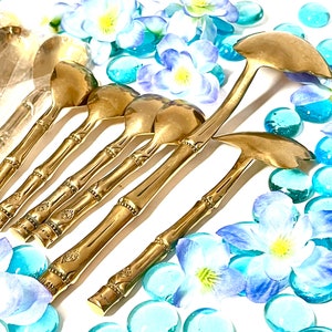 Puede incluir: Juego de seis cucharas de servir doradas con mangos de estilo bamb&uacute;. Las cucharas tienen un dise&ntilde;o decorativo y est&aacute;n dispuestas sobre una superficie blanca con flores azules y blancas.