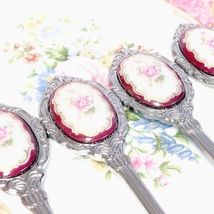 Pu&ograve; includere: Quattro specchietti da mano argentati con dettagli decorativi. Ogni specchio ha una cornice ovale rossa con un motivo floreale bianco e rosa.