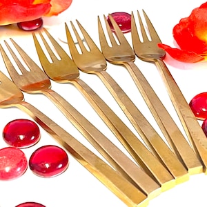 May include: Set of six gold-toned metal forks with square handles. The forks are arranged in a fan shape on a white surface with red glass stones.
