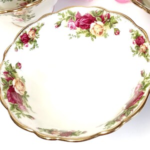 May include: A white ceramic bowl with a gold rim and a floral design featuring red, yellow, and green roses. The bowl has a scalloped edge.