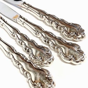 May include: Four silver dinner knives with ornate, detailed handles. The knives have a polished, shiny finish.