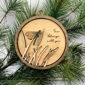 May include: A round wooden ornament with a laser-cut design of a dragonfly, cattails, and grass. The ornament has the text "I am always with you" inscribed on it.