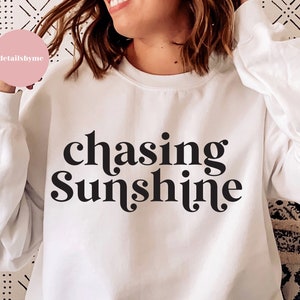 May include: White sweatshirt with black text that reads "chasing sunshine" in a retro font.