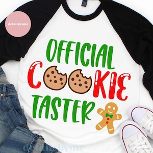 May include: A white and black baseball tee shirt with the text "OFFICIAL COOKIE TASTER" in green, red, and white. Two brown chocolate chip cookies with a bite taken out of them are on either side of the word "COOKIE". A gingerbread man with a green bow tie is below the text.