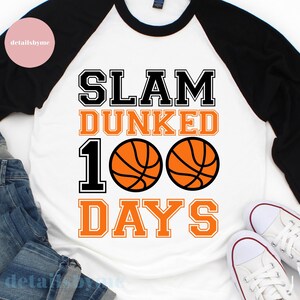 May include: A white and black baseball t-shirt with orange text that reads "Slam Dunked 100 Days". Two orange basketballs with black outlines are between the words "100" and "Days".