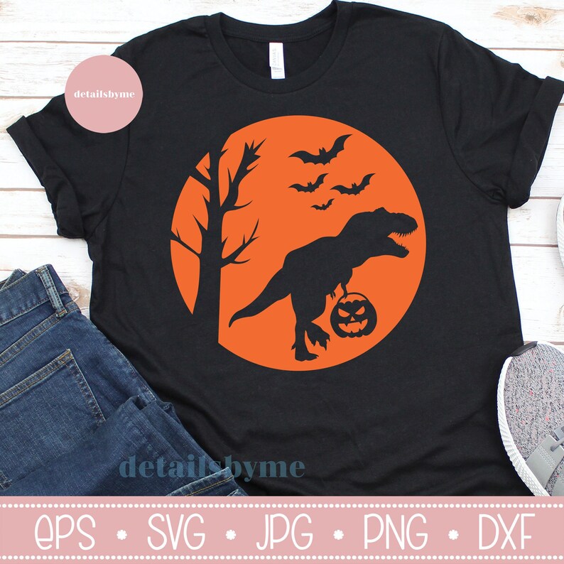 May include: Black t-shirt with an orange circle design featuring a silhouette of a dinosaur holding a jack-o'-lantern, a tree, and bats flying in the air. The text "detailsbyme" is at the bottom of the image.