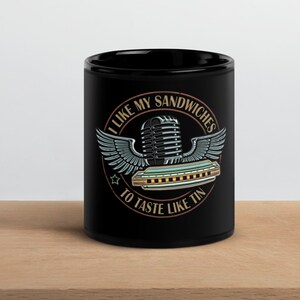 Op de afbeelding: Zwarte keramische mok met een afbeelding van een microfoon, mondharmonica en vleugels. De tekst op de mok luidt: "Ik vind mijn sandwiches lekker als ze naar tin smaken."