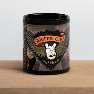 May include: Black ceramic mug with a rock and roll theme. The mug features the text "Horns Up!" above a skull with a hand gesture, wings, and the words "Pick Heads". A guitar graphic is on the side.