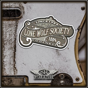 May include: A sticker featuring the words "LONE WOLF SOCIETY" in a vintage design, with "SINCE 1984" and "100% COOLNESS". Below is a smaller sticker reading "SR WEAR-A-TUDE". The background is a distressed, metallic surface.