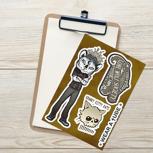 Puede incluir: Un portapapeles con un papel blanco en blanco y una hoja de pegatinas. Las pegatinas presentan ilustraciones de dibujos animados de un tigre, un gato calavera y una pancarta con el texto "LONE WOLF SOCIETY". Las pegatinas están sobre un fondo marrón.